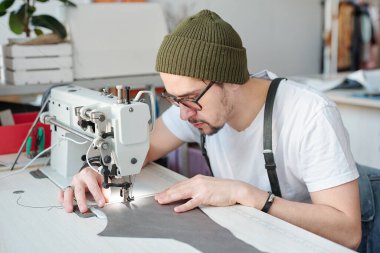 Ciddi genç erkek deri işçisi iş sırasında iğnenin altında deri parçasını tutarken elektrikli dikiş makinesinin üzerine eğiliyor.
