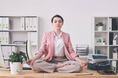 Pembe ceketli genç ruhani kadın lotus pozisyonunda masada oturmuş, ofiste meditasyon yapıyor.