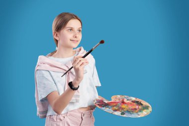 Tişörtlü, kot pantolonlu, elinde fırça ve renk paleti olan güzel genç bir öğrenci sanat okulunda resim yapmaya giderken...