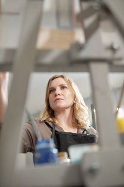 Önlüklü, sarışın, ciddi bir kadın ressam. Şövalenin önünde bitmemiş bir resim üzerinde çalışıyor.