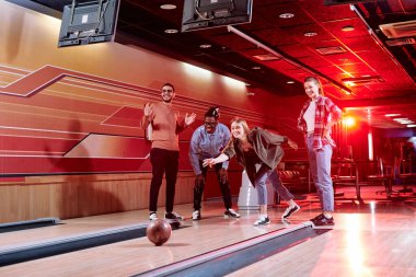 Çeşitli etnik kökenlere sahip bir grup neşeli arkadaş bovling salonunun yanında durup maç sırasında top atıyorlar.