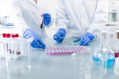 Beyaz yulaflı iki çağdaş bilim adamının eldivenli elleri laboratuarda masa başında dururken yeni bir dezenfektan ya da aşı deniyorlar.