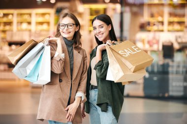 Alışveriş merkezinde kağıt torba taşıyan, günlük kıyafetler içindeki mutlu genç kadınların portresi.
