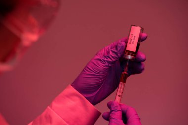 Kırmızı ışıkta Coronavirus hastalığına karşı şırıngayı aşıyla dolduran, eldiven giymiş, tanınmayan bir doktorun yakın çekimi.