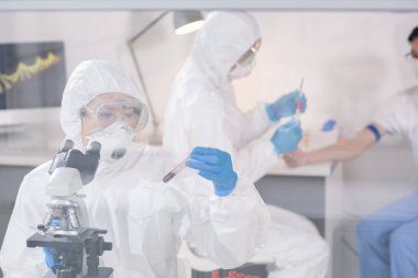 Koruyucu tulumlu genç bayan araştırmacı yeni aşı üzerinde çalışırken kan örneğiyle mataraya bakıyor.