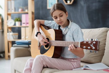 Rahat giyimli güzel kız kanepede oturup gitar çalmayı öğrenirken kendini karantina sırasında evde ders alıyor.