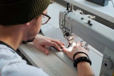 Genç erkek deri işçisi atölyede elektrikli dikiş makinesinin yanında otururken yeni deri cüzdan ya da başka bir şey yapıyor.