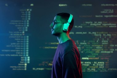 Kulaklık takan genç programcı ve günlük kıyafetlerin şifrelenmiş bilgilerle ekranın karşısında duruşu