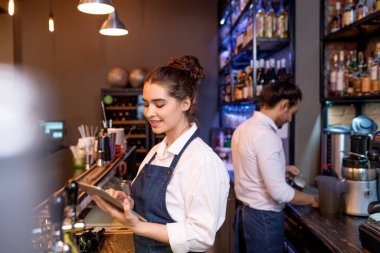 Meslektaşı arka plandaki konuklar için çay hazırlarken tabletli güzel garson kız internetten siparişleri karıştırıyor.