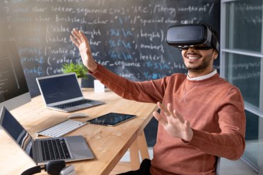 VR gözlük takan mutlu genç programcı dizüstü bilgisayarlarla oturup sanal ekrana bakıyor.