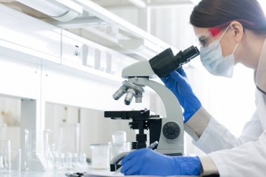 Beyazlı genç bir kadın, eldivenler, gözlük ve maske takarak mikroskop üzerinde eğilirken laboratuvarda koronavirüsün özelliklerini araştırıyor.