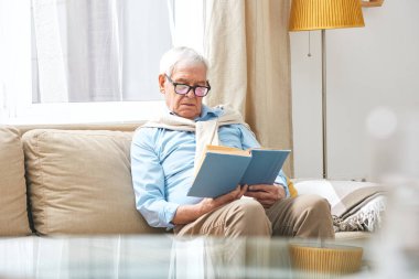 Kanepede oturup oturma odasında ilginç kitaplar okuyan, gözlüklü, ciddi yaşlı bir adam.