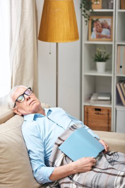 Gözlüklü yaşlı adam kanepenin altında oturuyor ve kitapla oturma odasında uyuyor.