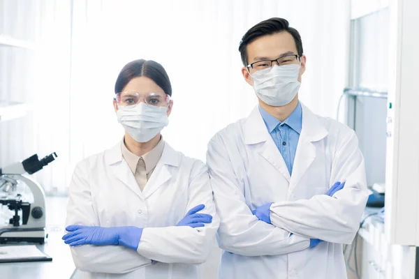 実験室でワクチンを研究しながらカメラの前に立つ白衣と手袋の二人の若い仮面の異文化科学者 — ストック写真