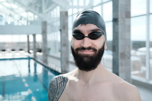 室内プールに立つ黒の帽子とゴーグルで満足した若い髭の水泳の肖像画 — ストック写真