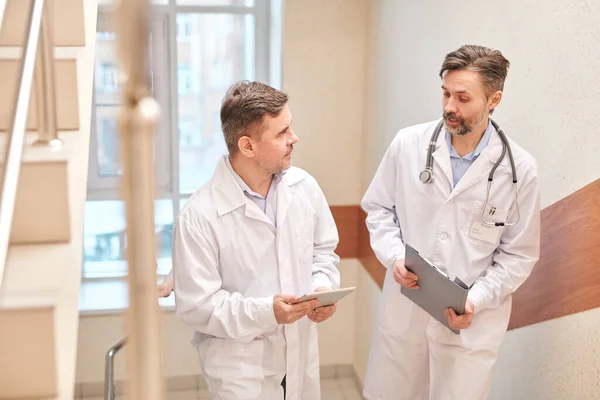 Mature doctors in lab coats moving up stairs and discussing diseases of patients