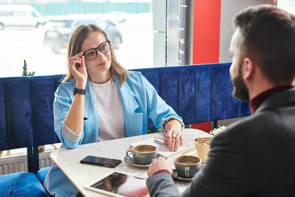 眼鏡を調整し 現代のカフェで会議で同僚とチャット魅力的な若いビジネスマンの女性 — ストック写真