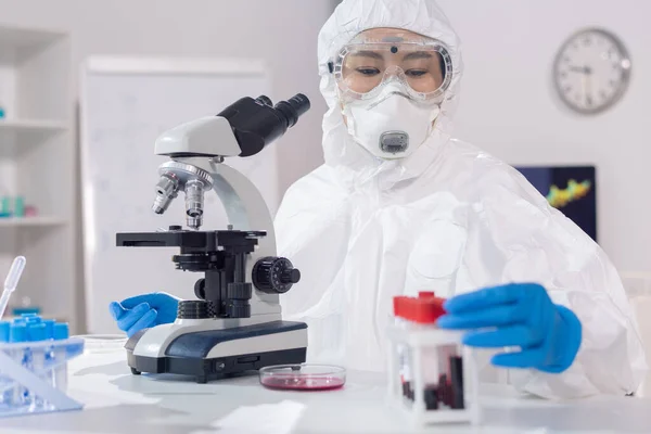 穿着工作服 面罩和防护眼镜的年轻妇女坐在实验室里 用显微镜检查血液样本 — 图库照片