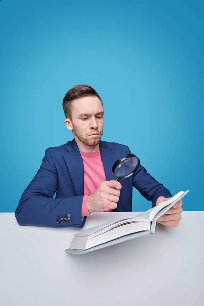 严肃的男性读者 带着放大镜坐在书桌旁 蓝色背景的书一页一页一页一页地翻阅 — 图库照片