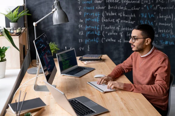 Young serious it-manager in eyeglasses sitting by table in front of computer monitor while decoding information in office