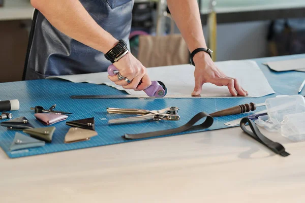 Hands of young leather worker using cutter while holding ruler on piece of white suede and cutting it on table