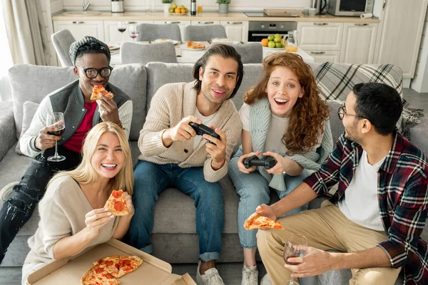 2人がホームパーティーでビデオゲームをしている間 若い異文化間の友人のグループはピザを楽しんでいます — ストック写真