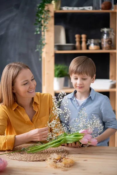 穿着衬衫的可爱男孩站在桌旁 帮助妈妈在厨房里摆弄郁金香花束和婴儿的呼吸 — 图库照片