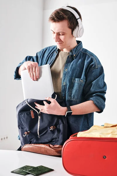 旅行中にサッチェルでワイヤレスヘッドフォンパッキングタブレットの陽気な若い学生 — ストック写真
