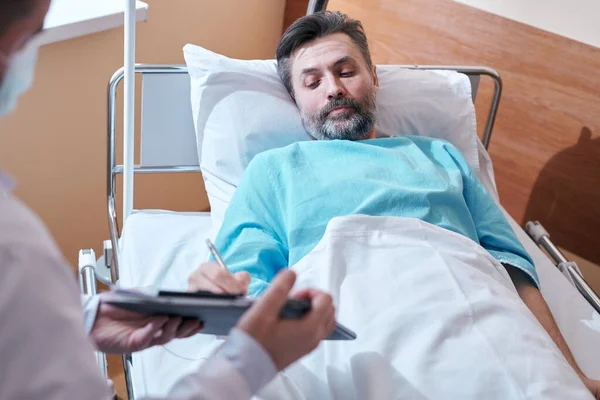 头戴大胡子 身穿医院长袍的病人躺在床上 在同意外科手术时签署医疗协议 — 图库照片