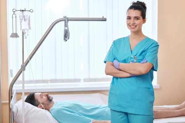 笑顔の若い看護師の肖像外科手袋に立って両腕で患者のベッドで病院 — ストック写真