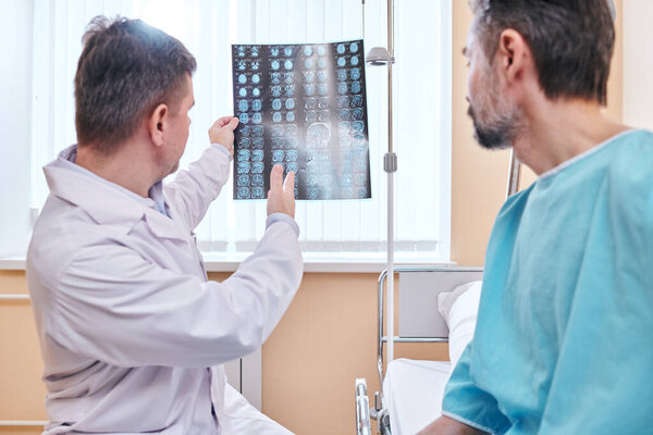 Experienced doctor in lab coat analyzing tomographic images while talking to patient in hospital