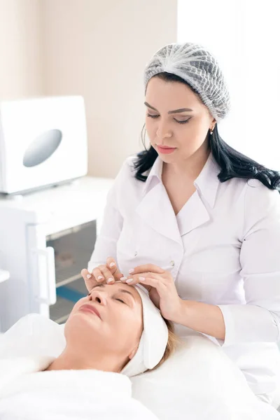 Beauty professional in surgical cap doing relaxing massage of face to mature client to smooth out wrinkles