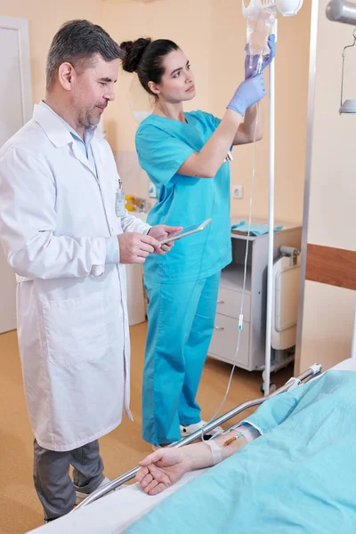 Młoda Pielęgniarka Przygotowująca Kroplówkę Podając Pacjentowi Dożylnie Dojrzały Lekarz Rozmawiający — Zdjęcie stockowe