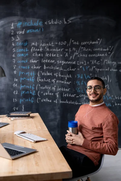 Young successful programmer with mug looking at you while sitting by desk on background of blackboard with written formula