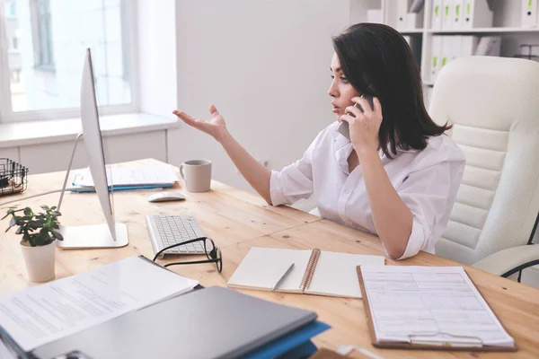 電話でビジネス上の問題を議論しながら オフィスのテーブルに座って コンピュータモニタを見て現代のビジネス女性 — ストック写真