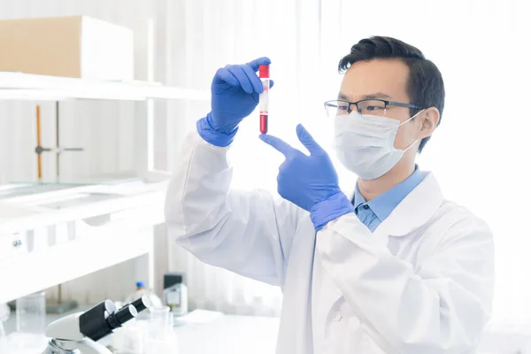 Young Chinese researcher in protective workwear looking at flask with red liquid substance while studying its characteristics in lab