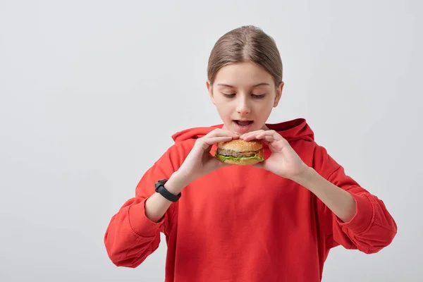 若いです女の子で赤パーカー保持食欲をそそりますハンバーガーによって彼女のオープン口の中でそれを食べますに対してホワイト背景で隔離 — ストック写真