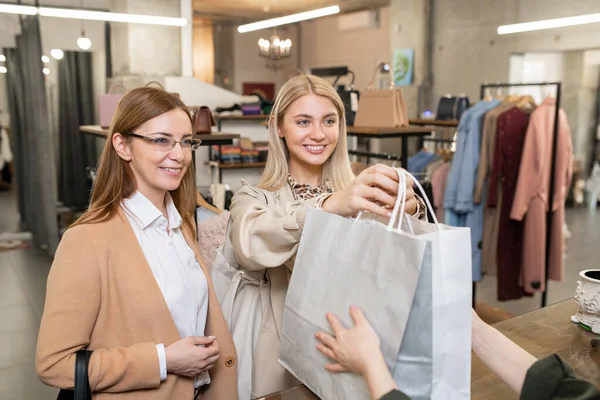 幸せな若い女性は ブティックで服を購入しながらカウンター越しにアシスタントを買い物に紙袋を渡す — ストック写真