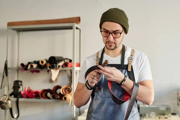 Professional craftsman in workwear holding red leather belt and wooden handtool while working over new item for client