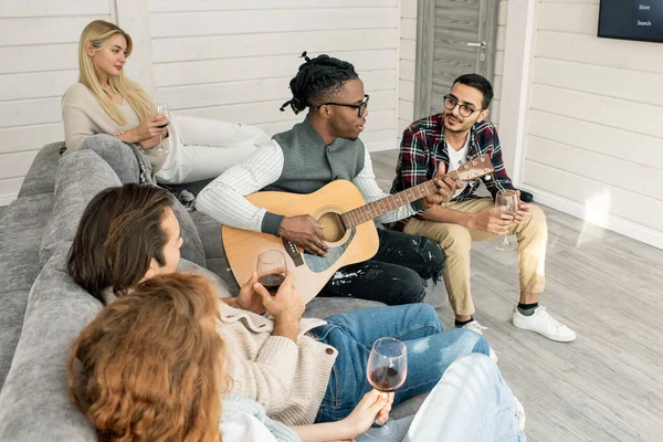 Joven Con Guitarra Cantando Para Sus Amigos Sentados Alrededor Sofá —  Fotos de Stock