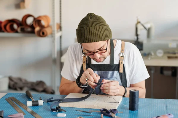车间用木板缝制新的黑色皮革制品时 年轻的创意皮革工人弯腰坐在桌子上 — 图库照片