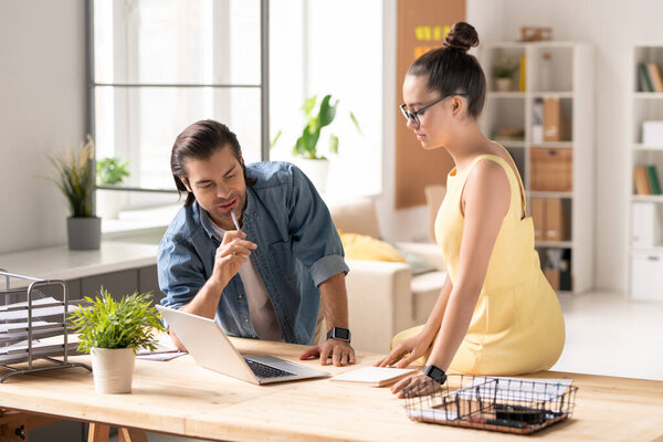 Young pensive businessman and his colleague looking at laptop display while discussing points of presentation before conference