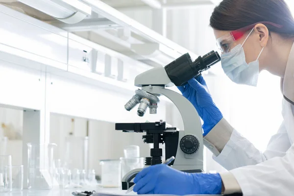 研究室でコロナウイルスの特徴を研究しながら アイウェア マスクの若い女性が顕微鏡に曲げ — ストック写真