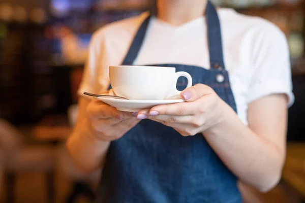 蓝色围裙上的年轻女服务员 为咖啡店或餐馆的顾客端着茶杯或卡布奇诺 — 图库照片