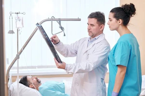 患者室で看護師と結果を議論しながら X線画像を指す研究室のコートで深刻な成熟した医師 — ストック写真