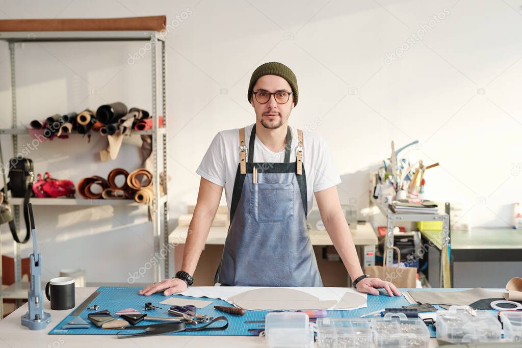 Young creative master in apron and beanie hat standing by table with working supplies in front of camera in workshop