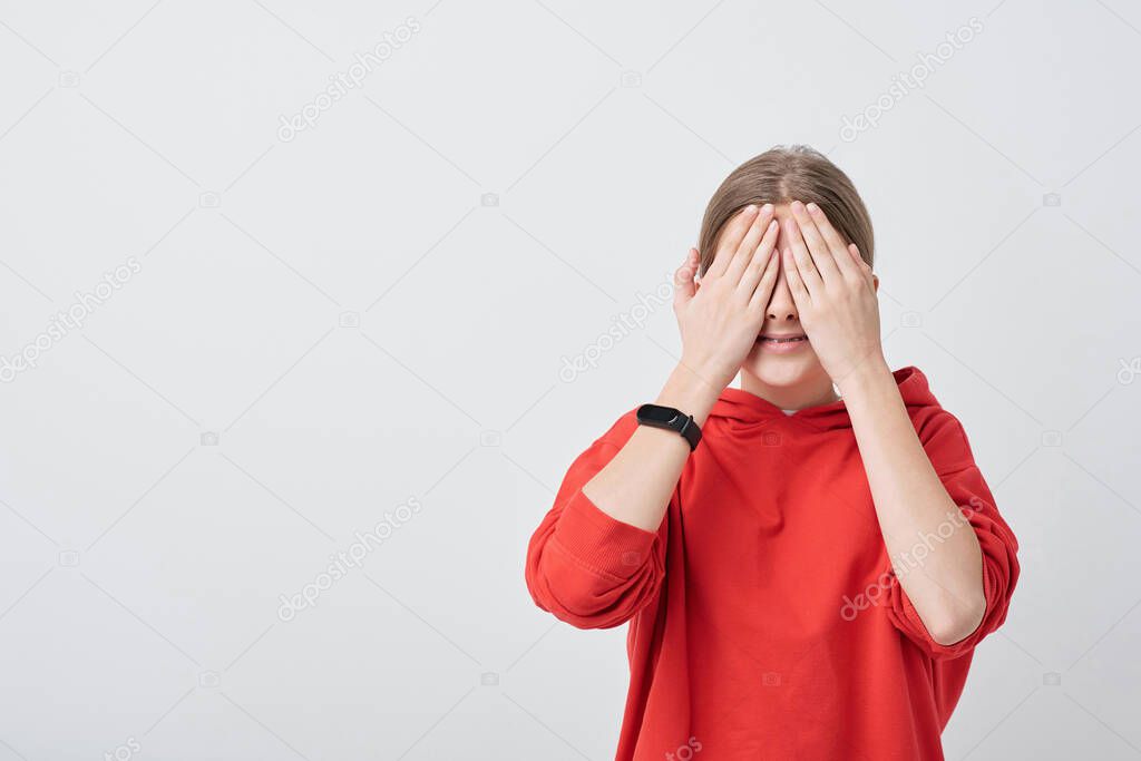 Smiling teenage girl in red hoodie covering her eyes by hands during hide-and-seek game while standing in front of camera