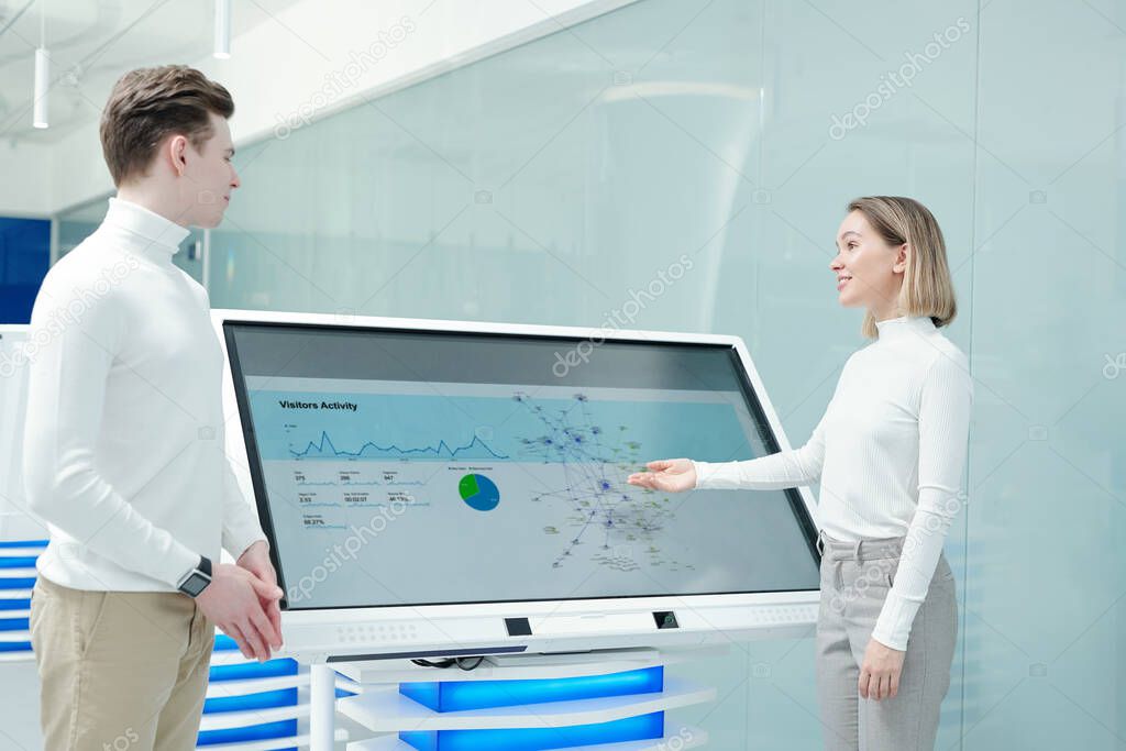 Positive friendly young woman pointing at interactive display and analyzing statistics of product distribution with colleague