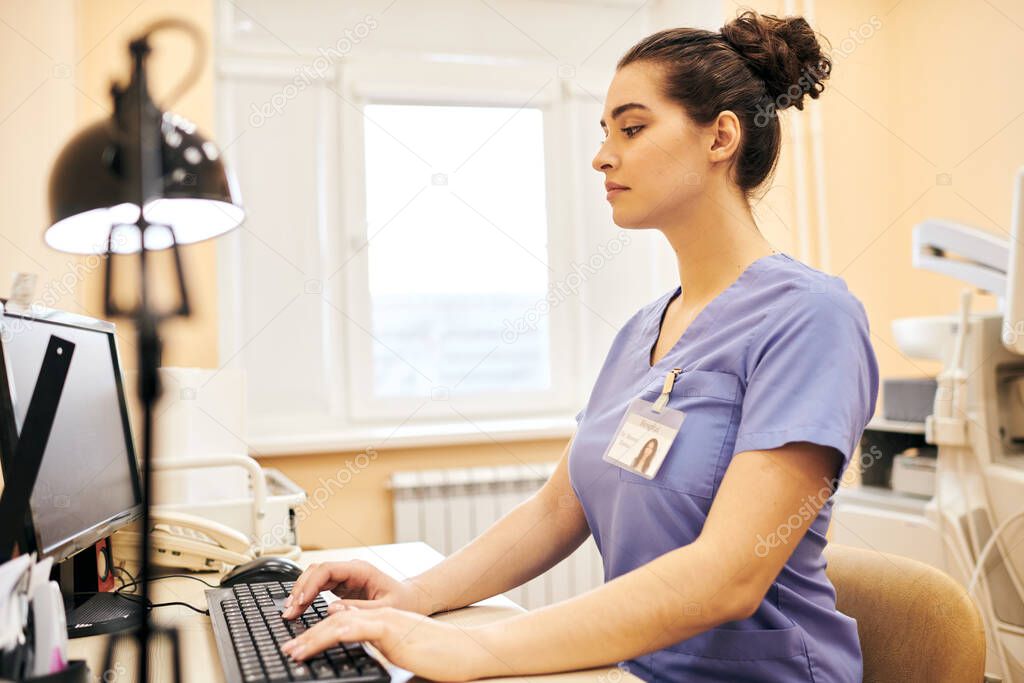 Concentrated young medical clinic employee in scrubs sitting at table and typing on computer keyboard