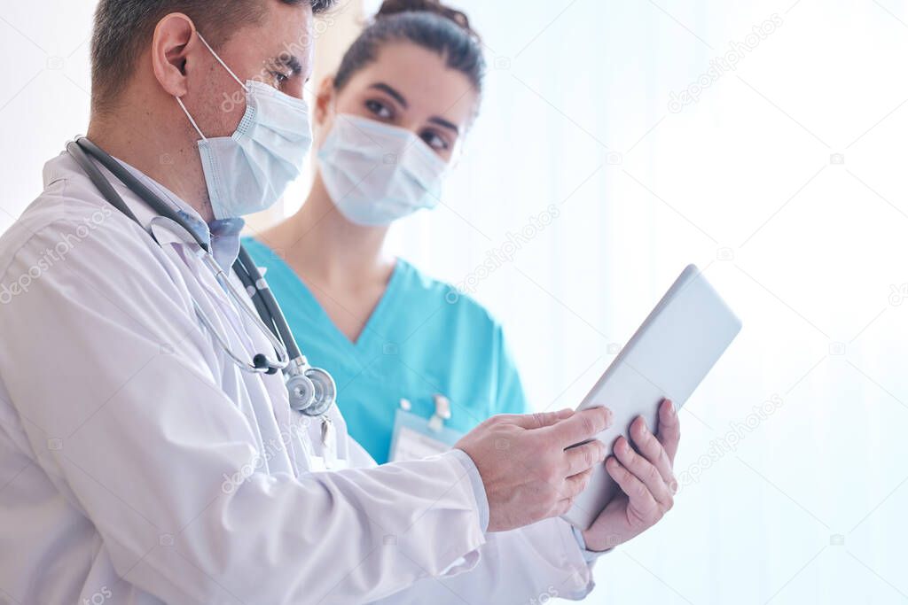 Doctor in mask and lab coat using digital tablet while discussing flu epidemic with colleague in clinic